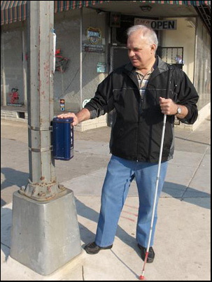 Man wearing jeans and black jacket holds a long cane in his left hand and stands on the sidewalk next to a pole. On the pole is a blue box about 4 by 4 inches on the top and 10 inches high.  The man has his right hand on top of the box (the box is about as high as his waist).